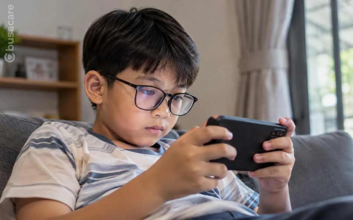 Anak berkacamata bermain ponsel di rumah dalam ilustrasi pentingnya menjaga kesehatan mata dan mencegah miopia pada anak sejak dini.