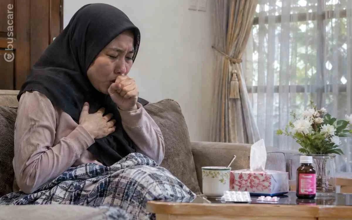 Seorang perempuan berhijab hitam mengalami batuk berkepanjangan sambil memegang dada saat beristirahat di rumah, ilustrasi pentingnya mengenali gejala batuk yang perlu diperiksakan ke dokter.